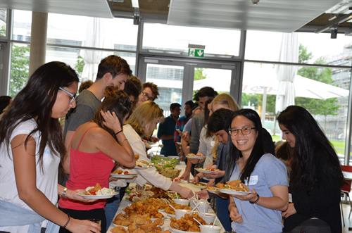 A group of students is standing at a long buffet table. A female student with glasses and black hair is smiling. She is holding a plate with food.
